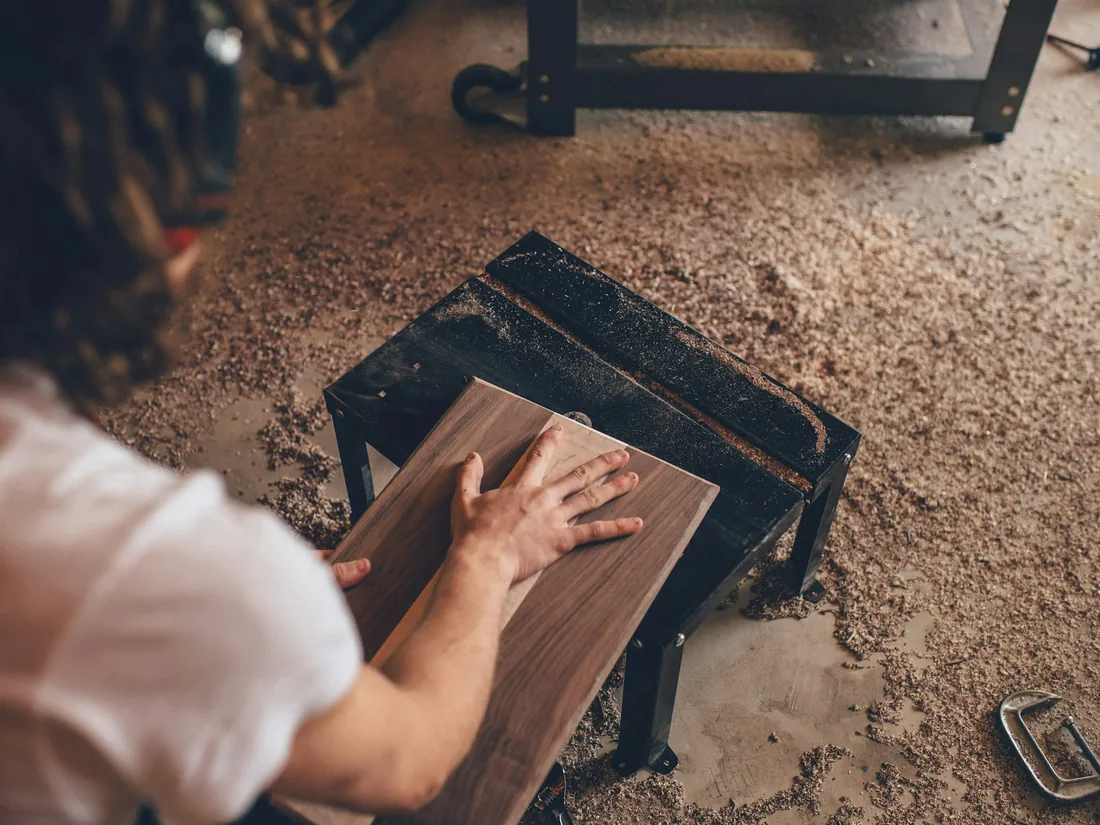 Manos sujetando una tabla de madera sobre un banco de taller cubierto de serrín y virutas